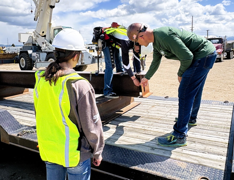 Three people inspecting with an iron girder that is lying on a trailer.